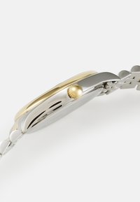 Montre en acier inoxydable avec une lunette dorée, un boîtier rond et une couronne dorée. Elle est dotée d'un bracelet argenté avec des maillons lisses.