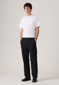 Man standing with hands in pockets, wearing a white t-shirt, black pants, black shoes, and a silver chain necklace against a plain background.