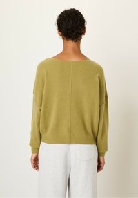 Pull en laine vert olive, surdimensionné avec des épaules tombantes et un large col. Texture douce et poignets côtelés. Associé à un pantalon de jogging gris.