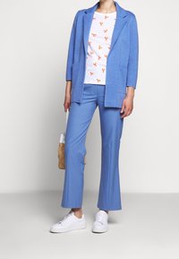 Ljusblå kavaj med fickor över en vit skjorta med orange hummertryck. Matchad med blå byxor och vita sneakers.