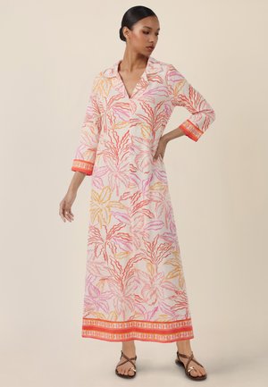 Frau steht mit Hand in der Hüfte, trägt ein langes weißes Kleid mit rosa, orangefarbenen Blattmustern und orangefarbenem Saum, kombiniert mit braunen Sandalen.