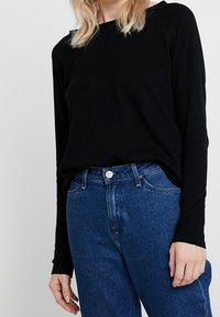 Svart långärmad topp i mjukt material, tillsammans med högmidjade blå denimjeans som har knäppning och framfickor.