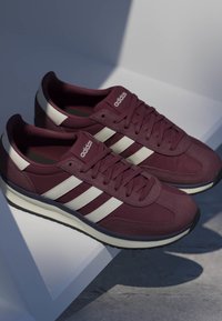 Baskets Adidas marron avec des bandes blanches et des semelles noires, lacées, reposant sur une surface blanche inclinée avec une ombre douce.