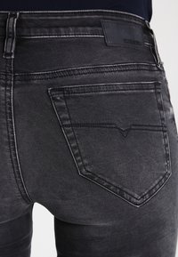 Los pantalones vaqueros de mezclilla negra tienen un ajuste ceñido con una textura desgastada. Los bolsillos traseros presentan un patrón de costura distintivo y una etiqueta de marca de cuero.