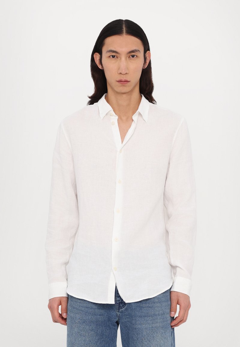 White linen long-sleeve shirt with a button-down collar, featuring a relaxed fit and subtle texture, worn with blue denim jeans.