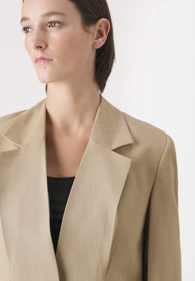 Beige maßgeschneiderter Blazer mit breiten Revers, glatter Textur und strukturiertem Schnitt, getragen über einem schwarzen Oberteil. Minimalistisches Design ohne sichtbare Beschläge.