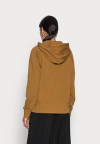 Hoodie marrone con cappuccio regolabile, polsini e orlo a coste, e una texture morbida. Indossato con una lunga gonna nera. Vista da dietro.