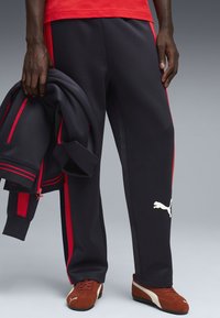 Σκούρες μπλε φόρμες με κόκκινες πλευρικές ρίγες, με λογότυπο PUMA. Κατασκευασμένες από απαλό ύφασμα, με χαλαρή εφαρμογή και χέρι που κρατάει ένα ταιριαστό μπουφάν.