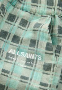 Grön och grå rutig tyg med en mjuk textur, med "ALLSAINTS"-märke och en etikett, stickad detaljering och en avslappnad design.