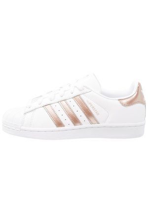 Biały sneaker Adidas Superstar z różowo-złotymi paskami i zakładką na pięcie, zamszowym noskiem i napisem "Superstar" po boku.