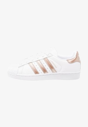 Biały sneaker Adidas Superstar z różowo-złotymi paskami i zakładką na pięcie, zamszowym noskiem i napisem "Superstar" po boku.