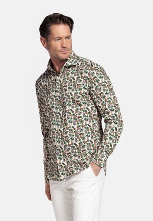 Man draagt een overhemd met knopen en lange mouwen met tropische dieren- en palmbomenprint, gecombineerd met een witte broek, staand met één hand in de broekzak.