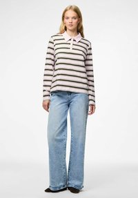 Jauna moteris stovi vilkėdama rožinės ir juodos spalvos dryžuotą ilgo rankovės polo marškinėlį, šviesiai mėlynus plačius džinsus ir juodus batus.