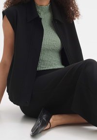 Czarna bezrękawnikowa kurtka nałożona na teksturowany zielony top, w połączeniu z czarnymi spodniami i czarnymi butami wsuwanymi. Gładkie materiały i luźny krój.