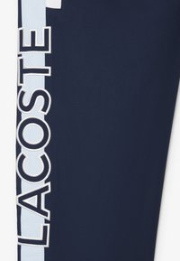 Marineblauwe broek met een lichtblauwe verticale streep en "LACOSTE" geborduurd in het wit aan de zijkant. Glad textiel, atletische pasvorm.