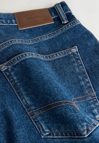 Gros plan de la poche arrière d'un jean en denim bleu avec couture orange et étiquette de marque en cuir marron sur la ceinture.