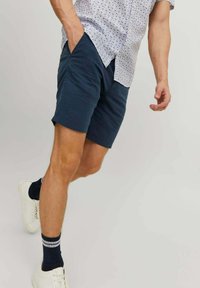 Șorturi bleumarin dintr-un material moale, cu o croială lejeră, tiv drept și buzunare laterale, purtate cu o cămașă cu un model și adidași albi.