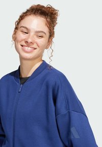 Sudadera azul con cremallera, cuello redondo, puños acanalados y un acento geométrico en la manga, hecha de una tela suave y texturizada.