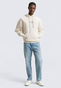 Béžová mikina s kapucí a šňůrkami, klokaní kapsou vpředu a potištěným textem. Kombinovaná se světle modrými džínami a bílými teniskami.