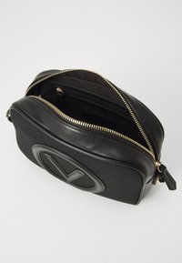 Μαύρη τσάντα με υφασμάτινη πρόσοψη, λείες δερμάτινες πλευρές, χρυσό φερμουάρ και έντονο ανάγλυφο λογότυπο στην μπροστινή όψη.