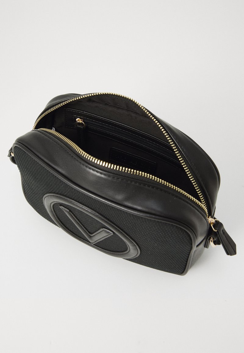 Sac noir avec une face texturée, des côtés en cuir lisse, une fermeture éclair dorée et un logo en relief bien visible à l'avant.