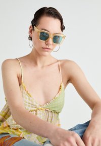 Gule gjennomsiktige solbriller med firkantede rammer, blå tonede linser og tynne stenger. Modellen har på seg en flerfarget, mønstret tanktop.