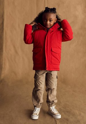 Chaqueta aislante roja con capucha forrada de pelo, que presenta dos bolsillos frontales. Combinada con pantalones cargo beige y zapatillas de color claro.