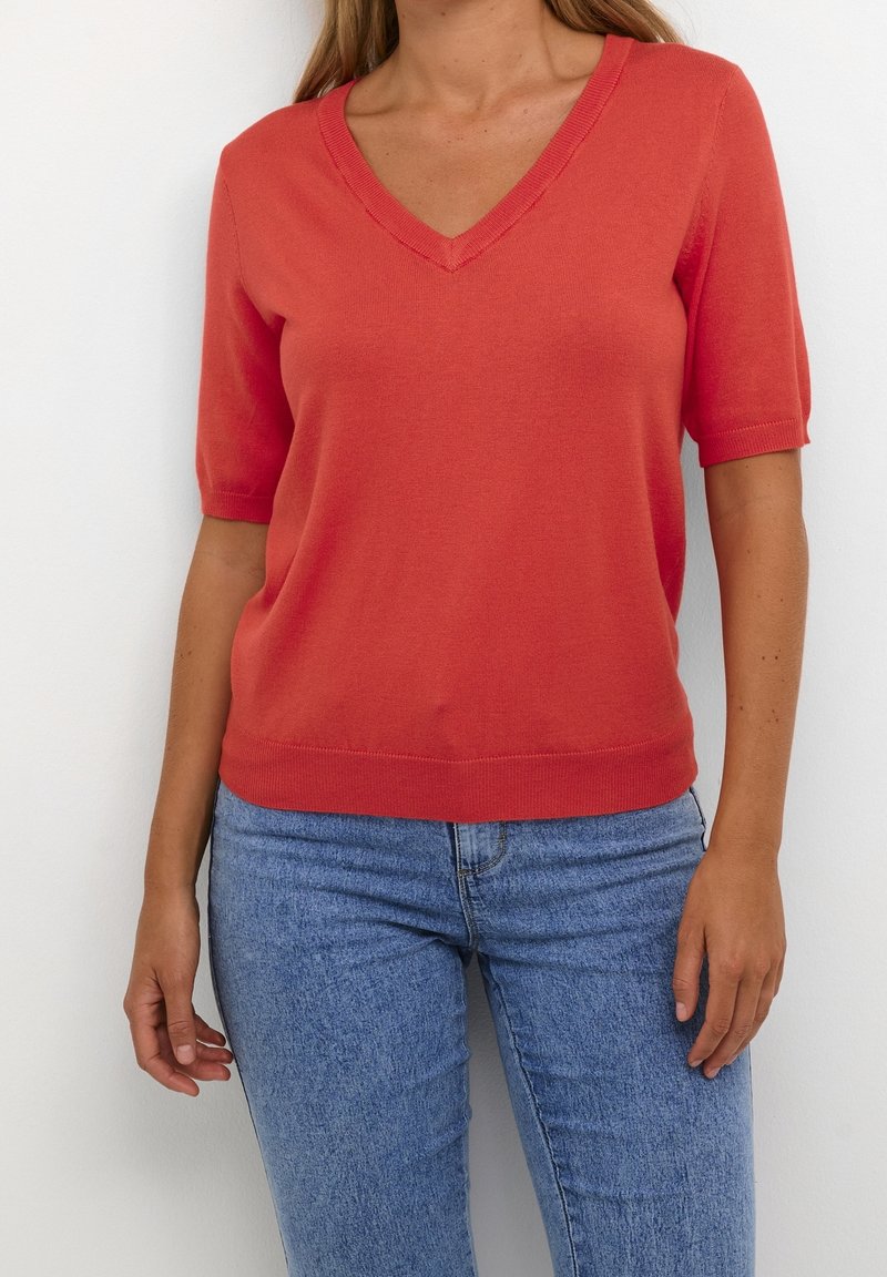 Coral short-sleeve V-neck knit top with ribbed neckline and hem, paired with blue high-waisted jeans, worn against a plain white background.