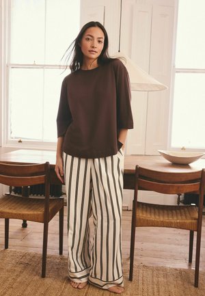 Woman in a brown loose top and white pants with black vertical stripes stands barefoot in a room with wooden chairs and a table.