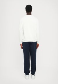 Suéter branco com gola redonda, mangas longas e bainha canelada, combinado com calças de jogging azul marinho e ténis brancos. Vista traseira mostrada.