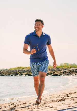 Blauwe poloshirt, gestreepte blauwe shorts, houdt een zonnebril vast, wandelt op een zandstrand nabij het water, casual zomeroutfit.