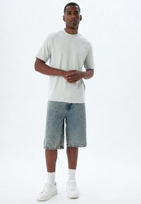 Lys grå kortærmet t-shirt sat sammen med slidte blå denimshorts, iført hvide sneakers og ankelsokker, stående på en ensfarvet baggrund.