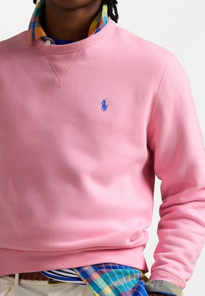 Lyserød sweatshirt med rund hals og blåt polo-logo. Båret over en flerfarvet ternet skjorte med et blåt og hvidt stribet mønster forneden.
