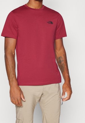 Camiseta roja de cuello redondo en algodón con mangas cortas y un logo negro en el lado izquierdo del pecho. Textura suave. Ajuste casual, combinada con pantalones beige.