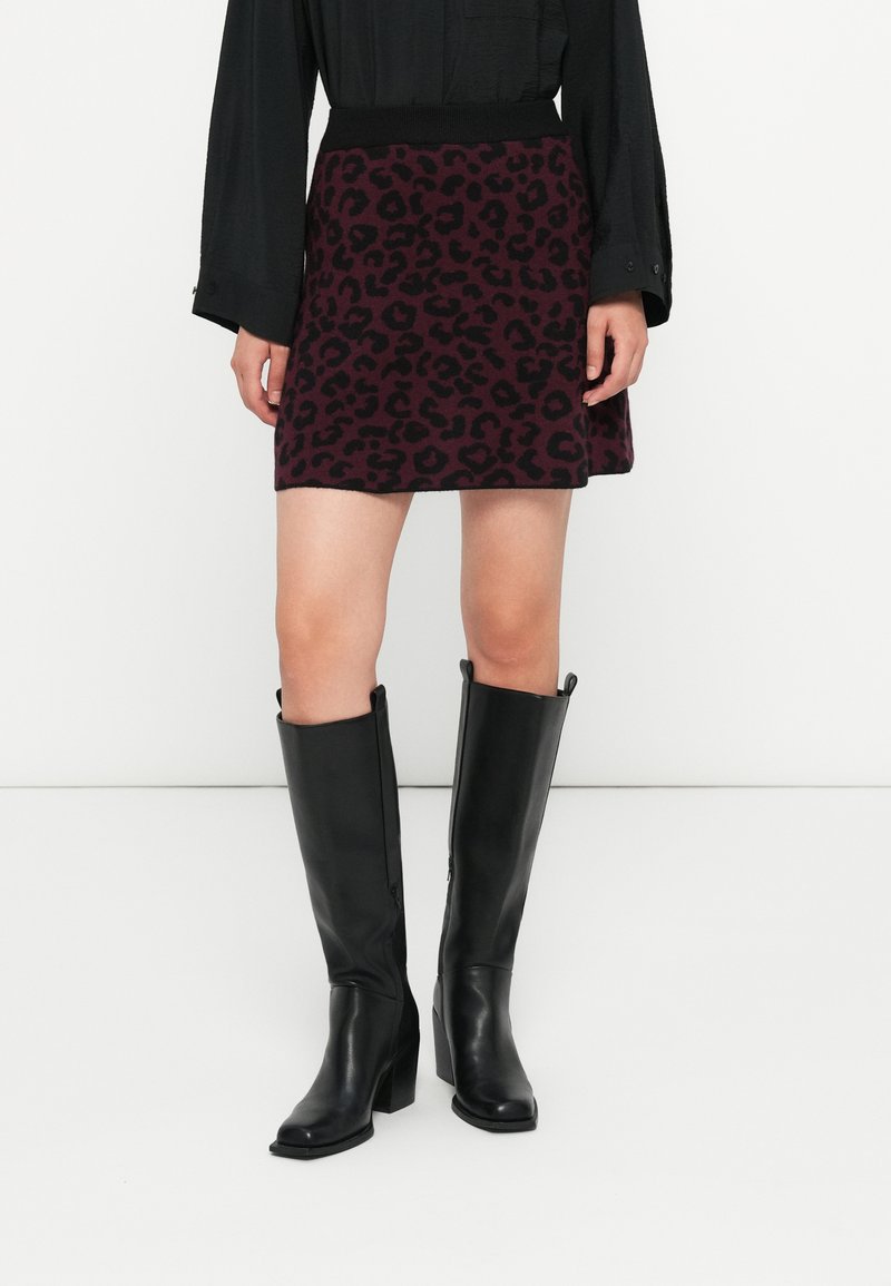 Mujer vestida con una blusa negra de manga larga, una falda mini de estampado de leopardo púrpura y botas de cuero negras de tacón alto hasta la rodilla, de pie contra un fondo blanco.