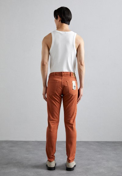 Rostfärgade byxor med slim fit, tillsammans med en vit linne. Byxorna har bakfickor och en etikett på midjan. Slät textur.