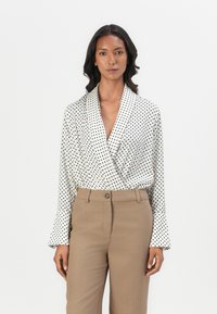 Hvit bluse med prikkete mønster, omslagsdesign og klokkelignende ermer, kombinert med brune skreddersydde bukser og kneppesteng.