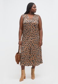 Γυναίκα με πλεξούδες που φοράει αμάνικο καφέ φόρεμα με μοτίβο λεοπάρ μέσης μήκους, καφέ σουέτ μπότακια αστραγάλου και κρατάει καφέ τσάντα.