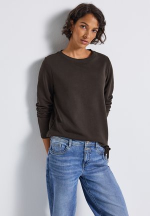 Femme aux cheveux courts et bouclés, portant un haut à manches longues sombre et un jean bleu, se tenant contre un mur blanc, les mains derrière le dos.