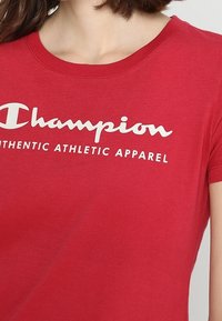 Červené bavlněné tričko s klasickým kulatým výstřihem a krátkými rukávy, s bílým potiskem: "Champion AUTHENTIC ATHLETIC APPAREL."