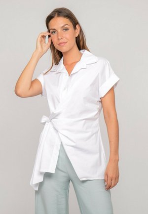 Femme portant un chemisier blanc à manches courtes avec ceinture nouée à la taille, associée à un pantalon ample bleu clair, posant devant un fond uni.