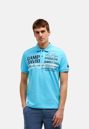 Mann steht mit den Händen in den Taschen, trägt ein hellblaues Poloshirt mit grafischem Text und dunkelblaue Shorts vor einem einfarbigen Hintergrund.
