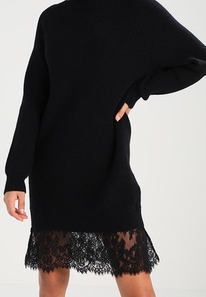 Czarna dzianinowa sukienka z długim rękawem, z przezroczystym, kwiatowym koronkowym dołem. Modelka pozuje, jedną rękę trzyma na biodrze, a drugą swobodnie opuszcza wzdłuż ciała.