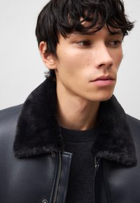 Giovane uomo con hair scuro che indossa una giacca di pelle nera con colletto in pelliccia sintetica e un maglione nero a maglia, che guarda verso destra.