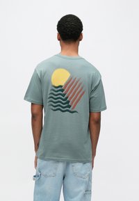 Zaļš kokvilnas T krekls ar lielu dzeltenu sauli, viļņiem un abstraktām svītrām tumši zaļā un brūnā krāsā uz muguras. Īsas piedurknes, brīvs siluets.