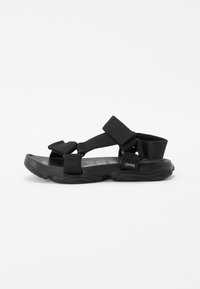Sorte svarte justerbare sandaler med tekstilremmer, en teksturert gummisåle og en litt konturert form. Merket er synlig på siden.