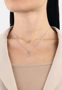 Srebrny naszyjnik z łańcuszka z zawieszką w kształcie półksiężyca ozdobioną małymi przezroczystymi kamieniami. Zawieszka ma błyszczące wykończenie; noszona na neutralnym topie.