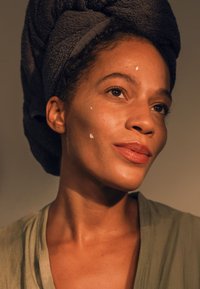 Woman with a dark towel on her head, wearing an olive green robe. She has light cream on her cheek and a subtle lip color. Soft lighting.