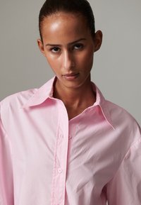 Šviesiai rožinė marškinėliai su sagutėmis, platus apykakle, ilgomis rankovėmis ir laisvo silueto. Lygus audinys su šiek tiek raukšlelių prie sagų.