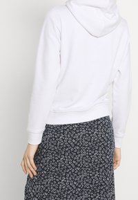 Bílá mikina s kapucí na stažení, s žebrovanými manžetami a lemem. Spárováno s černou sukní zdobenou malými bílými květinovými vzory.